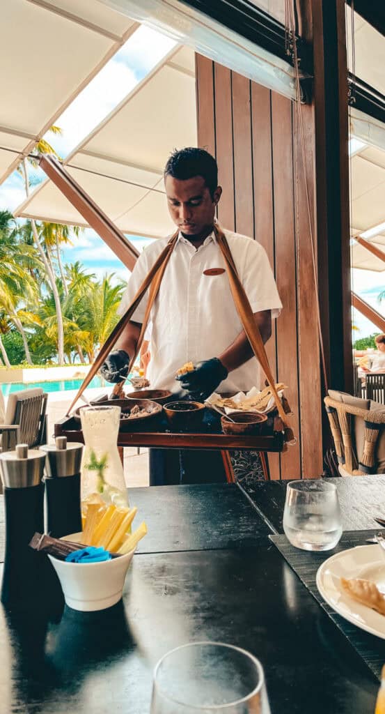 A chef preparing food at a live breakfast station at a Maldivian resort, focusing intently on spreading condiments on a piece of flatbread. He wears a white uniform with a name badge and is under a wooden canopy, with tropical palm trees visible in the background, enhancing the dining experience with a local touch.