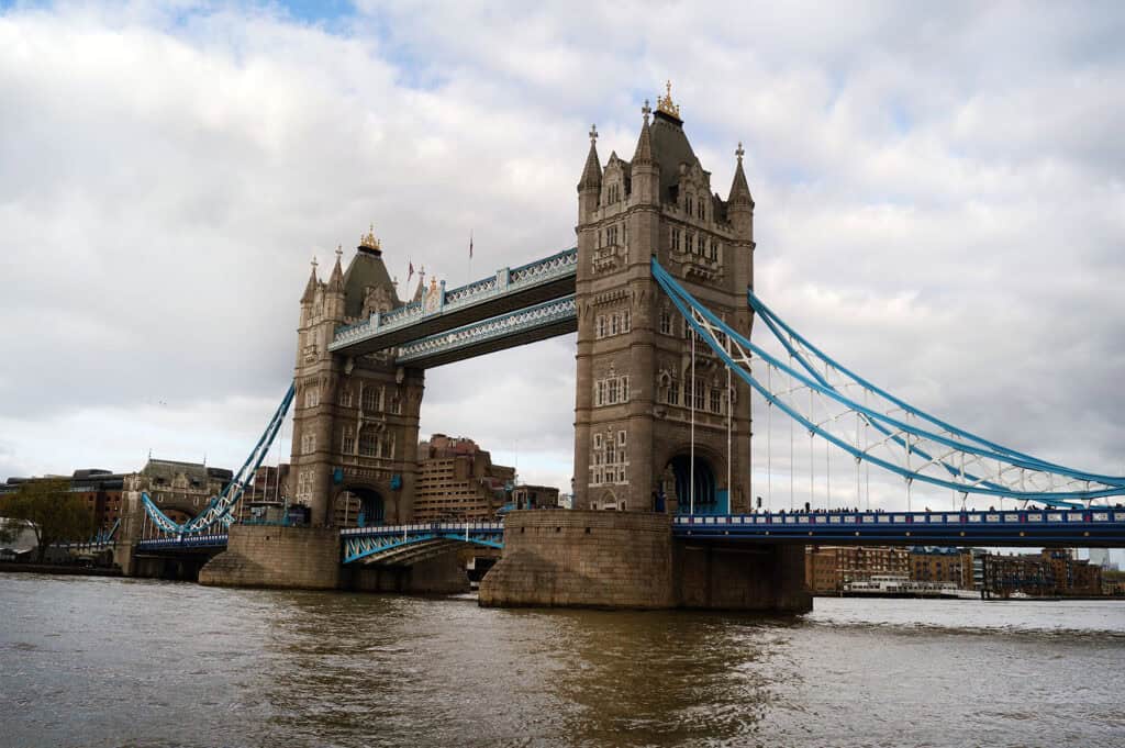 A view of London's iconic Tower Bridge spanning the River Thames, featuring its two Gothic-style towers connected by upper walkways and blue suspension elements. The cloudy sky and surrounding cityscape add to the historic and urban charm of this famous landmark.