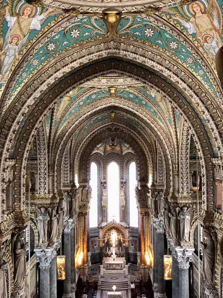 An intricate and ornate interior of a cathedral in Lyon, France, featuring richly decorated arches with gold accents and colorful patterns. Statues of angels and saints adorn the columns, leading the eye towards the grand altar illuminated by soft lighting. Stained glass windows cast a serene glow over the elaborate architectural details.