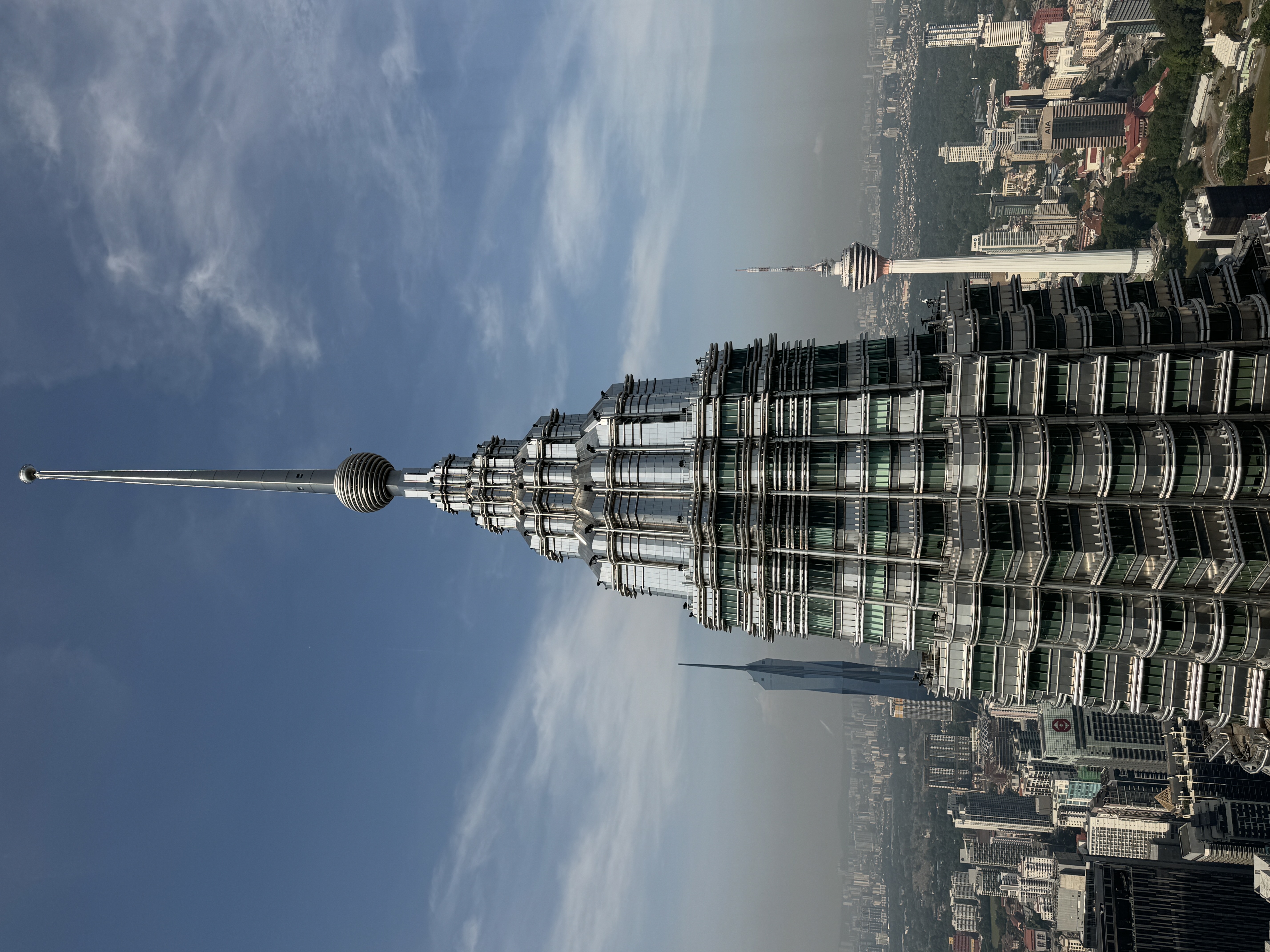 A close-up view of one of the Petronas Towers in Kuala Lumpur, Malaysia, highlighting its intricate architectural design and spire against a clear blue sky. The Kuala Lumpur Tower is visible in the background, along with the sprawling cityscape below. This image captures the modern and iconic skyline of the Malaysian capital.
