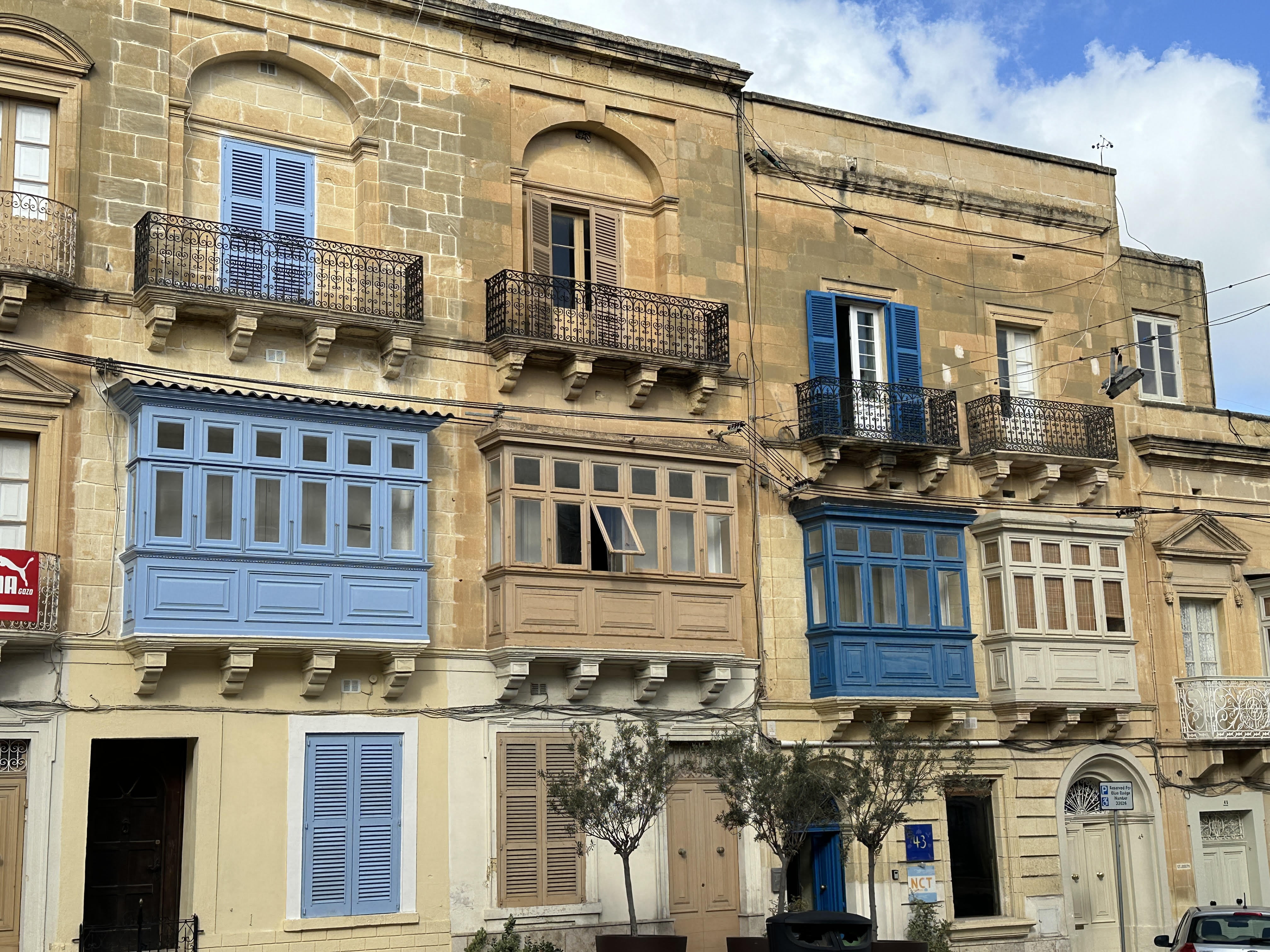 A charming street view in Malta, showcasing traditional Maltese balconies painted in pastel shades of blue, beige, and white. The stone buildings feature ornate wrought-iron railings and shutters, adding to the picturesque and historic ambiance. The bright sky and a few trees in the foreground complete this quaint urban scene.