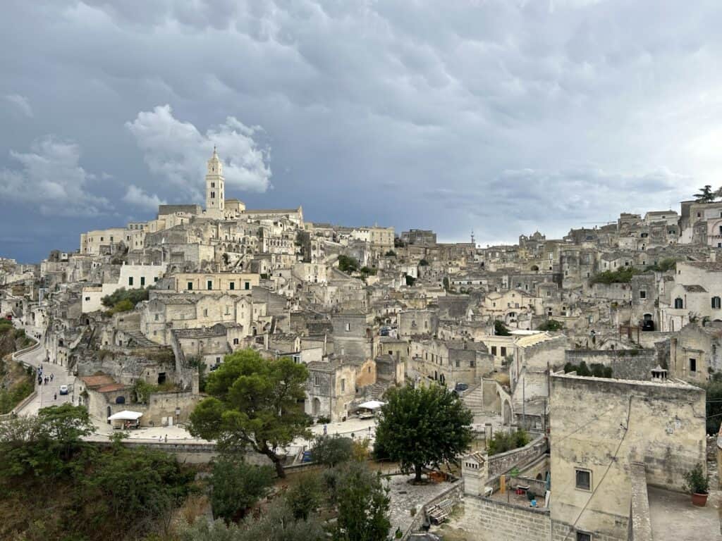A panoramic view of Matera, Italy, showcasing its historic stone buildings and narrow streets under a cloudy sky. The city's iconic cathedral with its tall bell tower stands prominently on the hilltop, surrounded by densely packed ancient structures. Lush greenery and scattered trees add a touch of nature to the urban landscape.