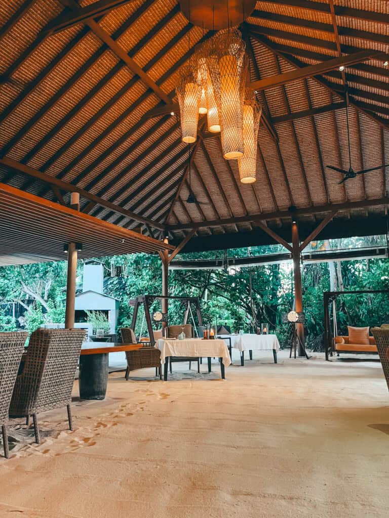 An outdoor dining area with sandy floors and a high thatched roof, featuring wicker chairs and tables set for a cozy meal, illuminated by warm hanging lights and surrounded by lush greenery.