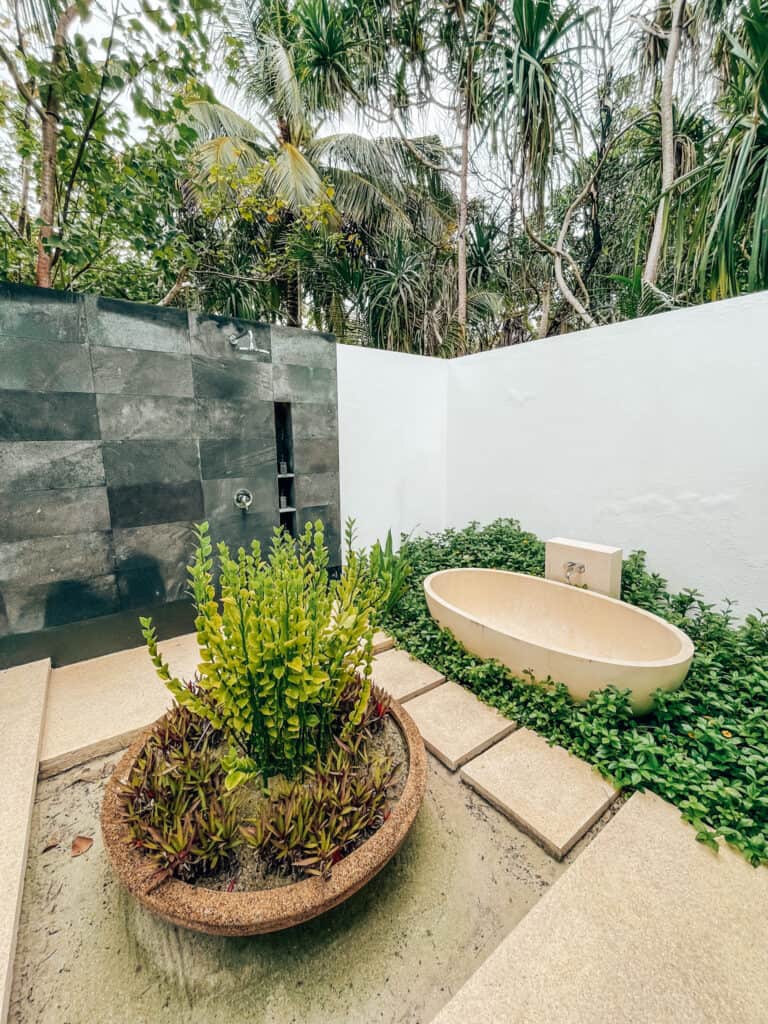 Outdoor bathroom setting surrounded by dense tropical foliage, featuring a stone wall and a white oval bathtub nestled among greenery and potted plants. The open-air design captures a serene and private spa-like atmosphere, inviting relaxation in a natural environment