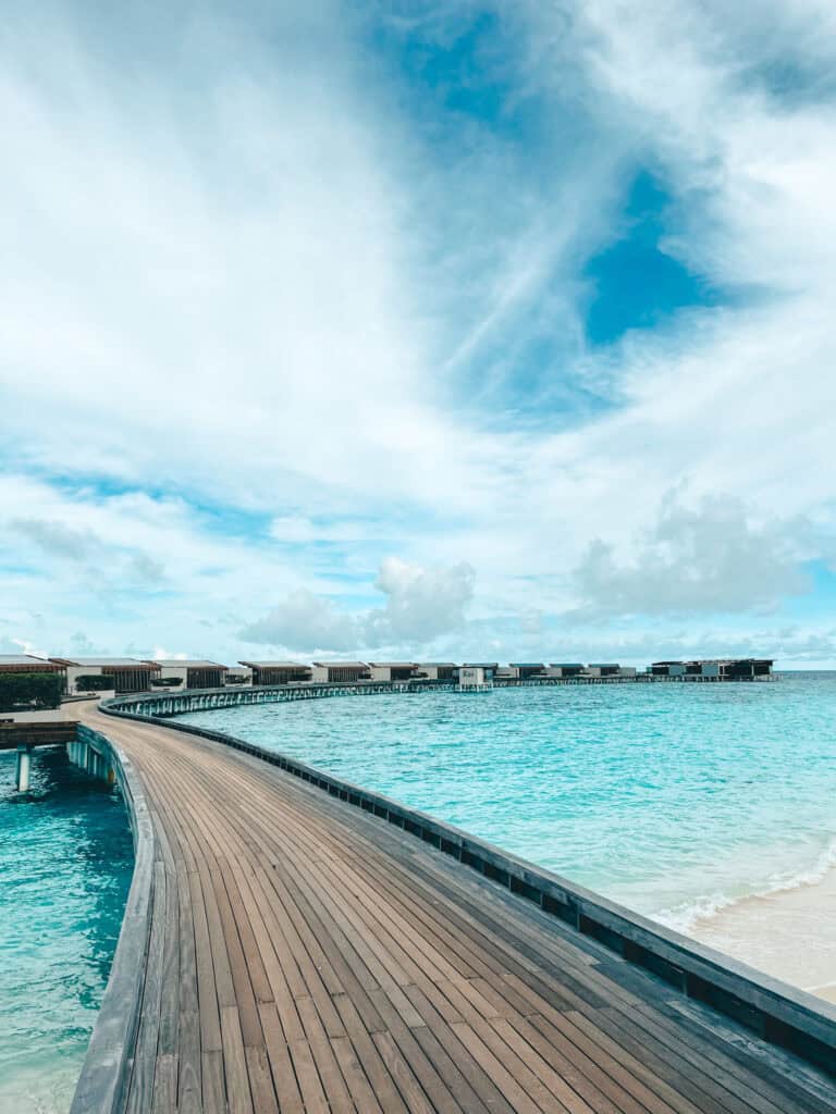 A long wooden pier extending over clear turquoise water, leading to a series of overwater villas under a bright blue sky with scattered clouds. The scene is tranquil and picturesque, perfect for a luxurious tropical retreat.