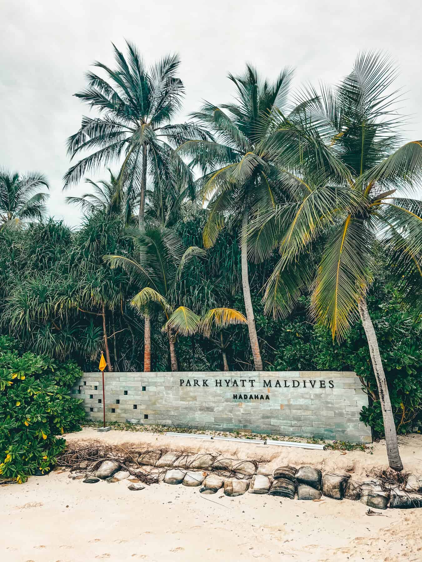 Entrance sign of Park Hyatt Maldives Hadahaa set against a lush background of palm trees on a sandy beach. The sign, built into a low stone wall, is surrounded by vibrant green foliage under a cloudy sky, embodying a tropical and welcoming atmosphere