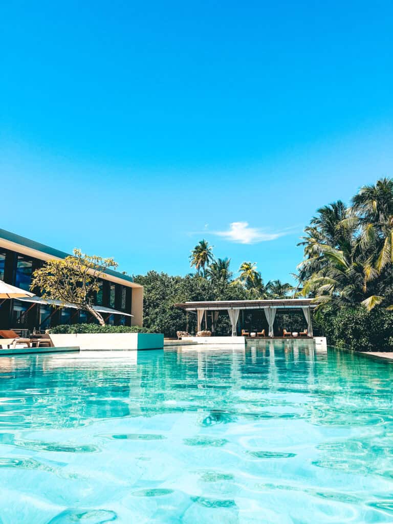 Inviting view of a large resort swimming pool with crystal clear turquoise water, reflecting the bright blue sky above. The pool is bordered by modern architecture and lush tropical vegetation, featuring a pool bar with shaded seating areas, providing a perfect spot for relaxation and leisure in a serene setting.