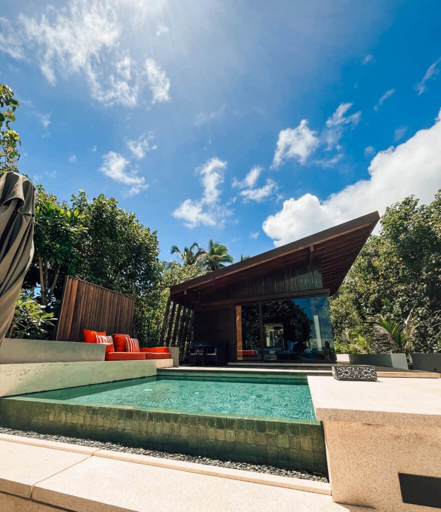 Modern villa exterior showcasing a private swimming pool surrounded by a wooden deck and lush tropical vegetation. The architectural design features a wide glass facade and an angled dark wooden roof, with a bright sky and fluffy clouds above, offering a perfect blend of natural beauty and contemporary luxury