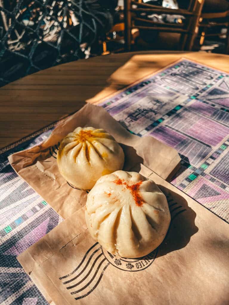 Two pork bao buns with a light dusting of spices sit on a piece of brown paper, placed on a table with a colorful, patterned tablecloth. The scene is bathed in warm sunlight, casting shadows and highlighting the soft, fluffy texture of the buns.