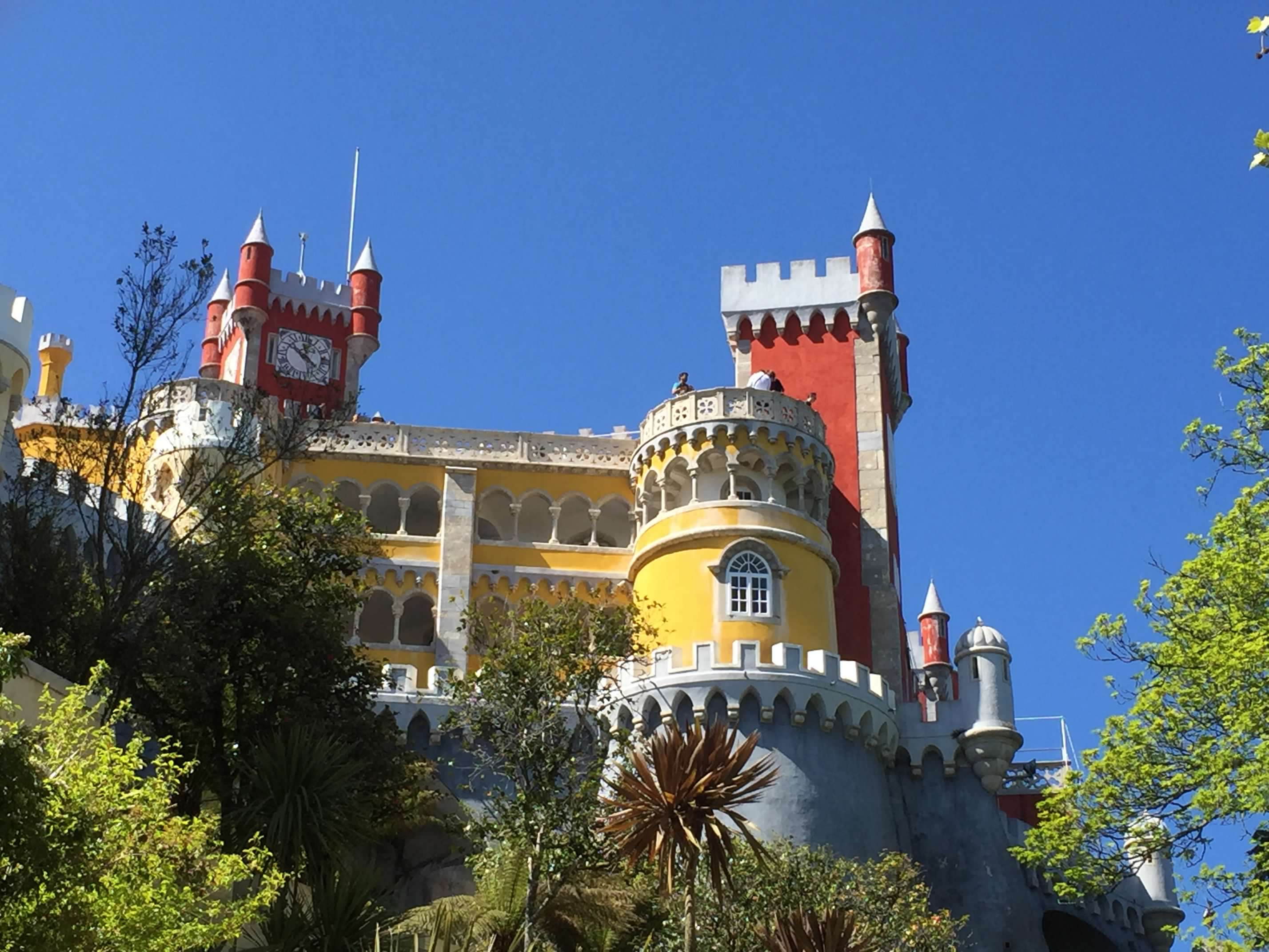 A vibrant view of Pena Palace in Sintra, Portugal, displaying its striking red and yellow towers under a clear blue sky. The palace's whimsical architecture features ornate balconies and crenellated walls, surrounded by lush greenery. This iconic landmark stands out against the natural landscape, creating a fairy-tale-like scene.