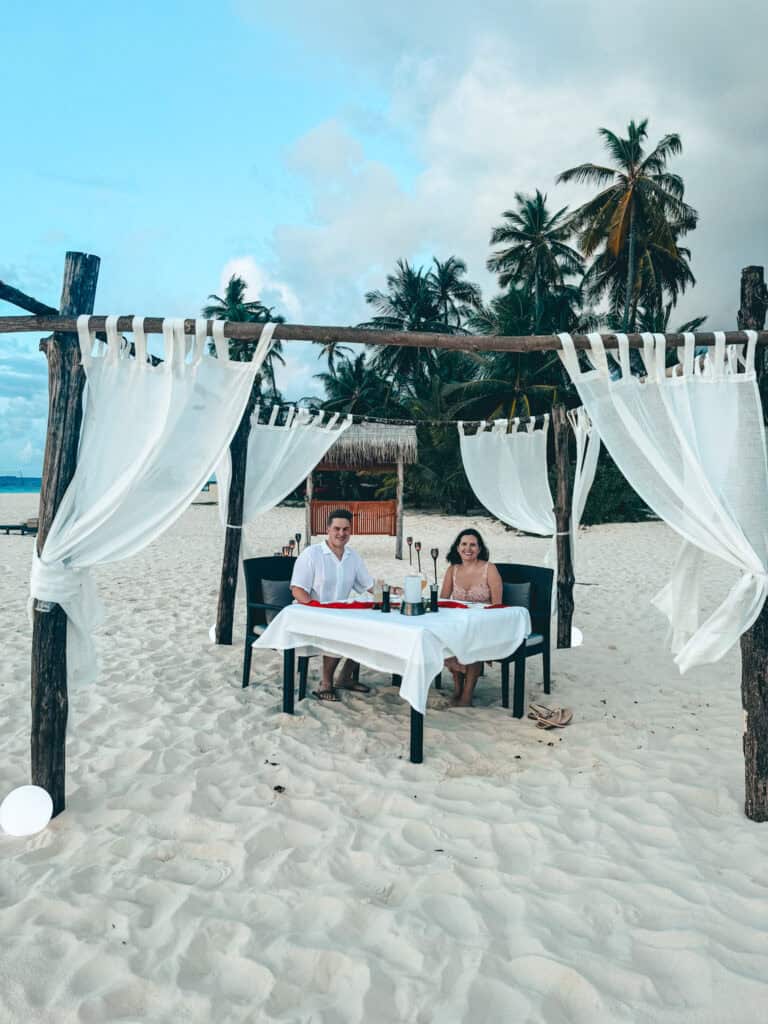  couple enjoying a private dinner on the beach, seated at a table under a wooden canopy with flowing white curtains, set against a backdrop of palm trees and a clear blue sky.