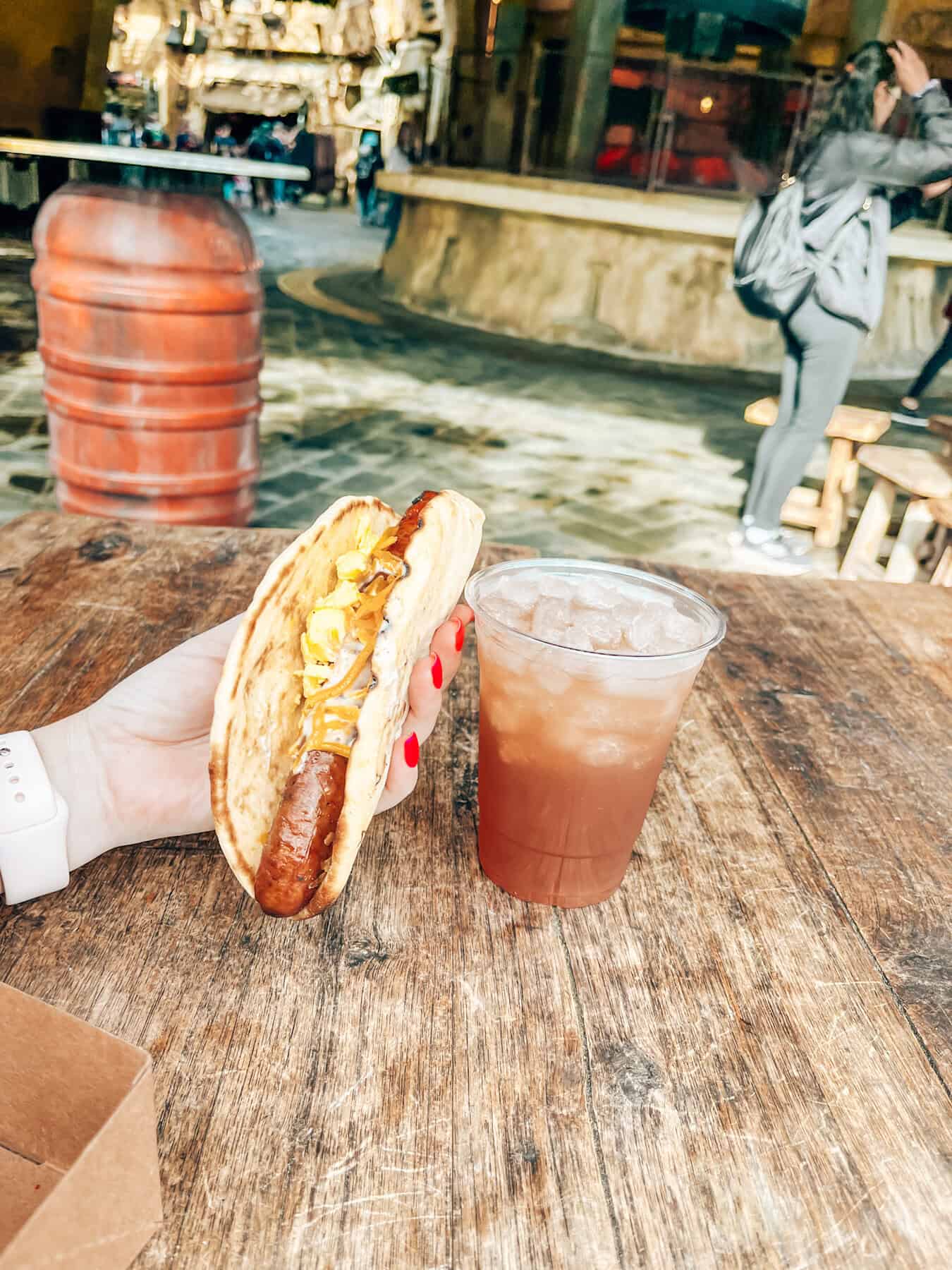 A hand with red-painted nails holds a Ronto Wrap, consisting of grilled sausage, slaw, and sauce wrapped in flatbread, over a rustic wooden table. Next to the wrap is a plastic cup filled with an iced beverage, with a blurred, themed setting in the background.