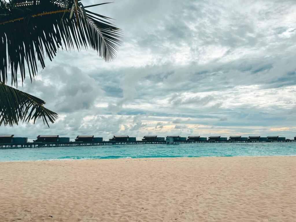 View of a sandy beach at the Park Hyatt Maldives showing a row of overwater villas extending into the azure sea under a dramatic cloudy sky. The beach is pristine and deserted, emphasizing the peaceful and exclusive atmosphere of the resort.