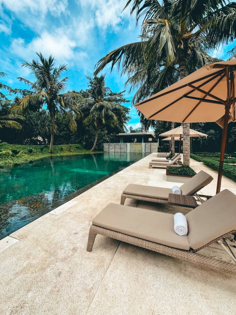 Luxurious poolside area at a resort spa featuring beige sun loungers under a large tan umbrella, beside a calm, reflective water pool. The setting is surrounded by lush tropical trees and a clear blue sky, offering a tranquil and inviting atmosphere for relaxation.