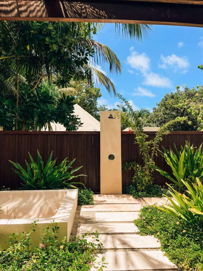 Outdoor spa shower area in a private garden enclosed by a high wooden fence and lush greenery. The pathway leads to an open-air shower with a stone wall, enhancing the serene and natural spa experience amidst vibrant tropical plants.