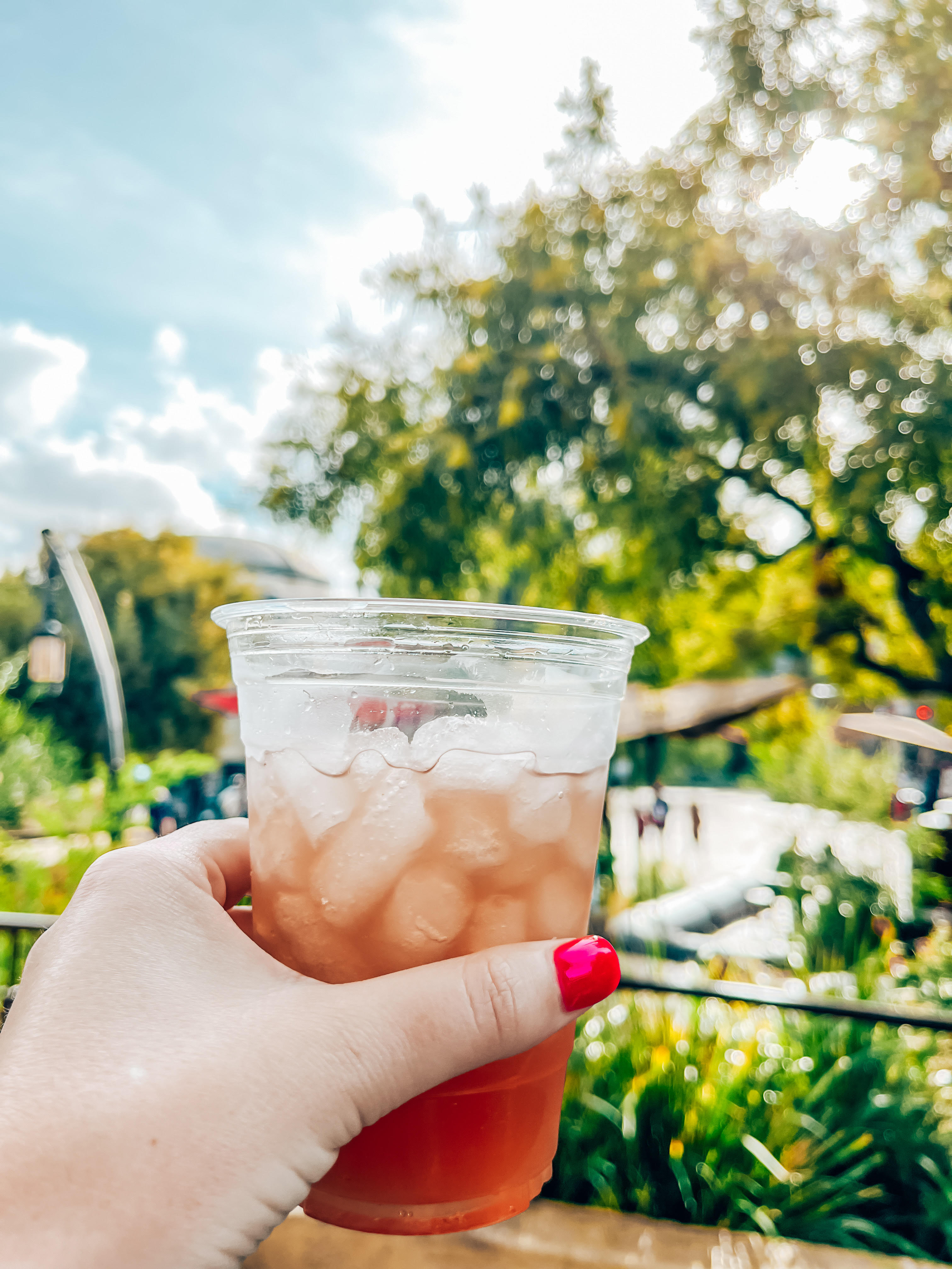 A hand with pink-painted nails holds a plastic cup filled with a reddish-orange iced beverage, known as a Tatooine Sunset, against a backdrop of lush greenery and a bright, sunny sky. The scene captures a refreshing moment in a picturesque outdoor setting.