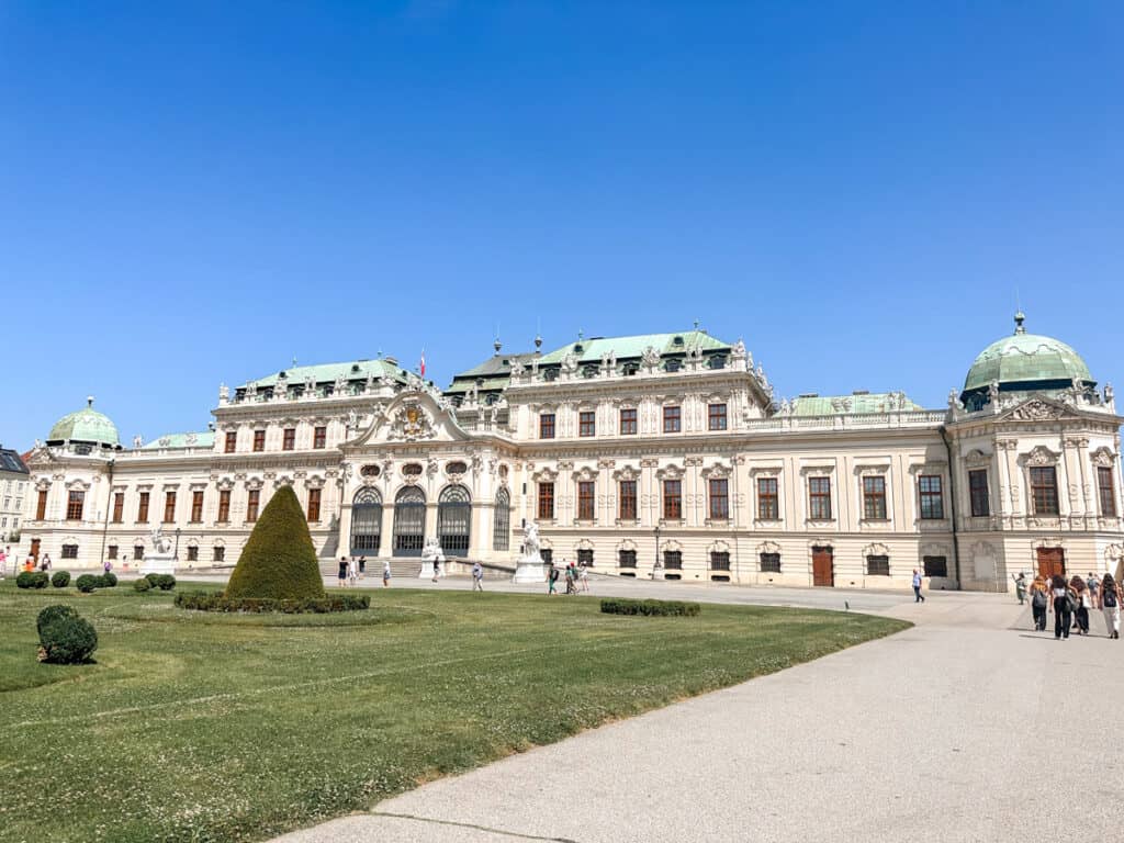 A grand Baroque-style palace with green copper roofs and ornate architectural details stands under a clear blue sky in Vienna, Austria. The well-manicured garden in the foreground features a conical topiary and neatly trimmed shrubs, with a few visitors walking along the paths.