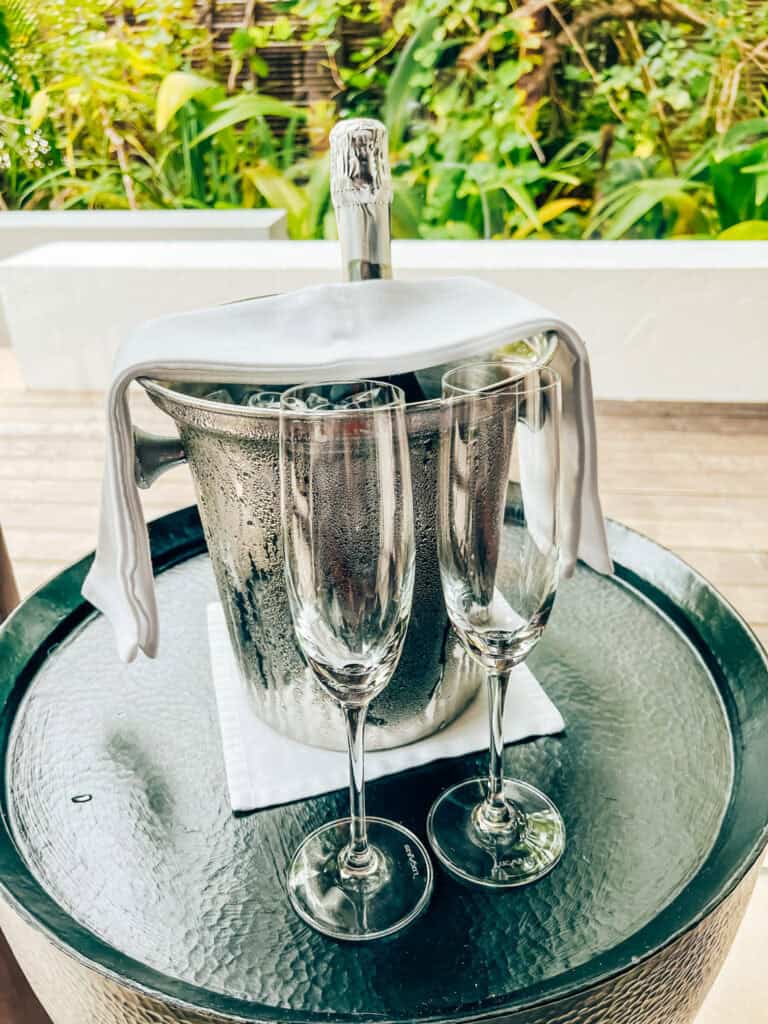 Welcome setup at a resort with a bottle of champagne chilling in a metal ice bucket, accompanied by two empty champagne glasses on a glass-topped round table. The setting is outdoors, with lush greenery in the background, creating a celebratory and inviting atmosphere