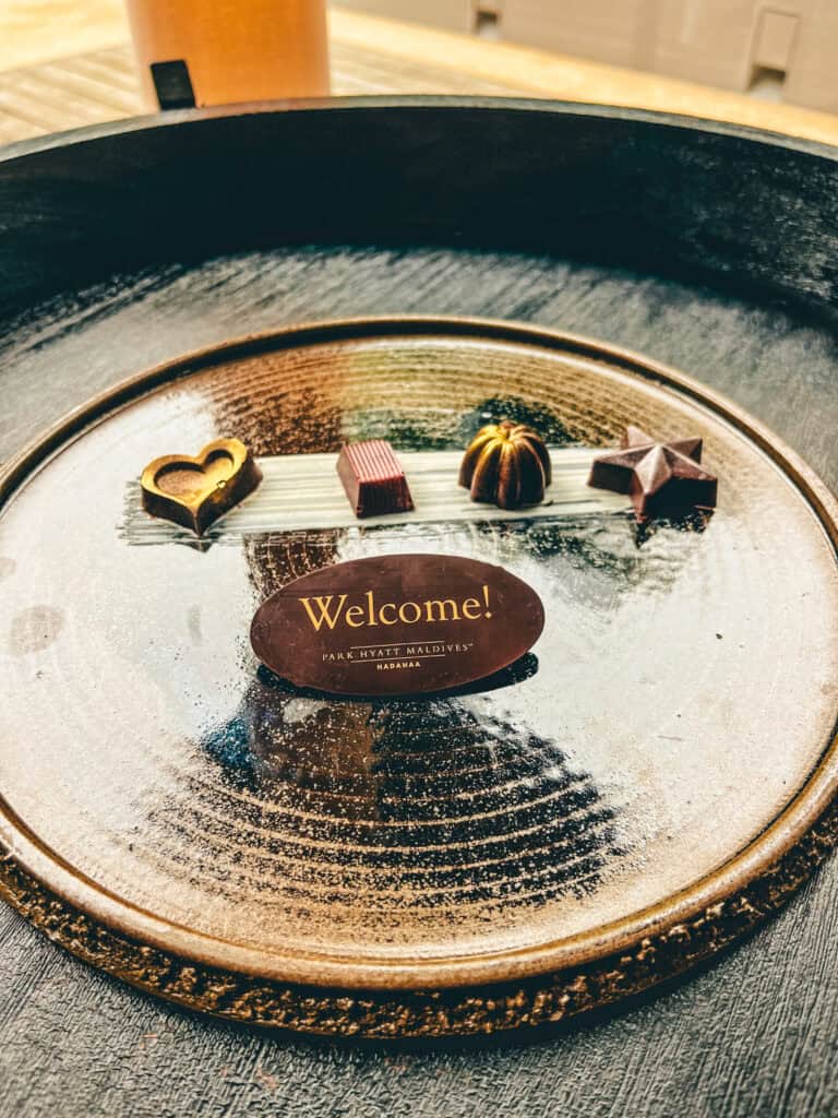 Elegant welcome treat featuring a selection of artisan chocolates arranged on a ceramic plate with a 'Welcome! Park Hyatt Maldives Hadahaa' chocolate placard. The chocolates, including heart-shaped and assorted geometric designs, are presented on a textured plate, adding a luxurious touch to the greeting