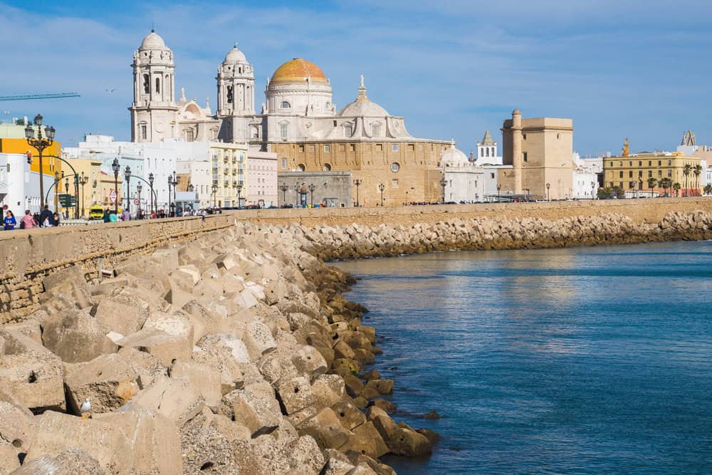 A scenic view of Cádiz, Spain, featuring the grand Cádiz Cathedral with its golden dome and twin bell towers rising above the city's skyline. The cathedral and surrounding historic buildings are seen from a rocky sea wall, with the blue waters of the bay contrasting against the bright, clear sky. Visitors stroll along the waterfront, enjoying the sunny weather and picturesque setting.