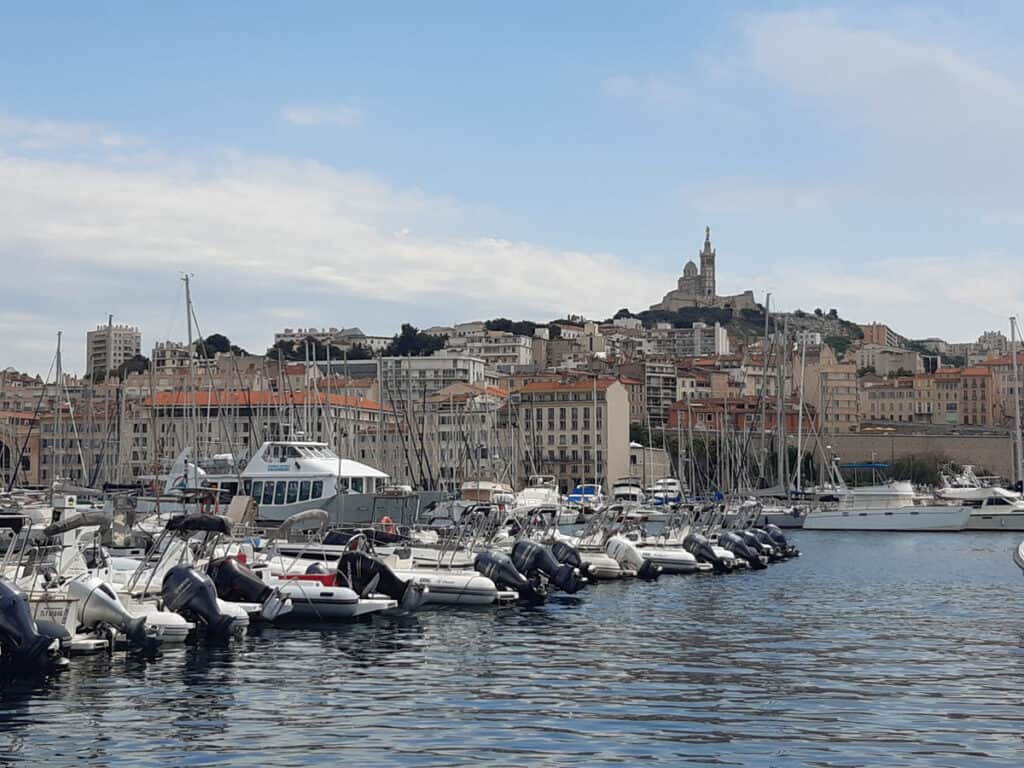 A view of the Vieux-Port (Old Port) of Marseille, France, filled with numerous boats and yachts moored in the marina. The city's skyline, featuring terracotta-roofed buildings, rises behind the harbor, with the iconic Basilique Notre-Dame de la Garde perched on a hilltop in the distance. The scene is set under a partly cloudy sky, adding to the charm of this historic Mediterranean port city.