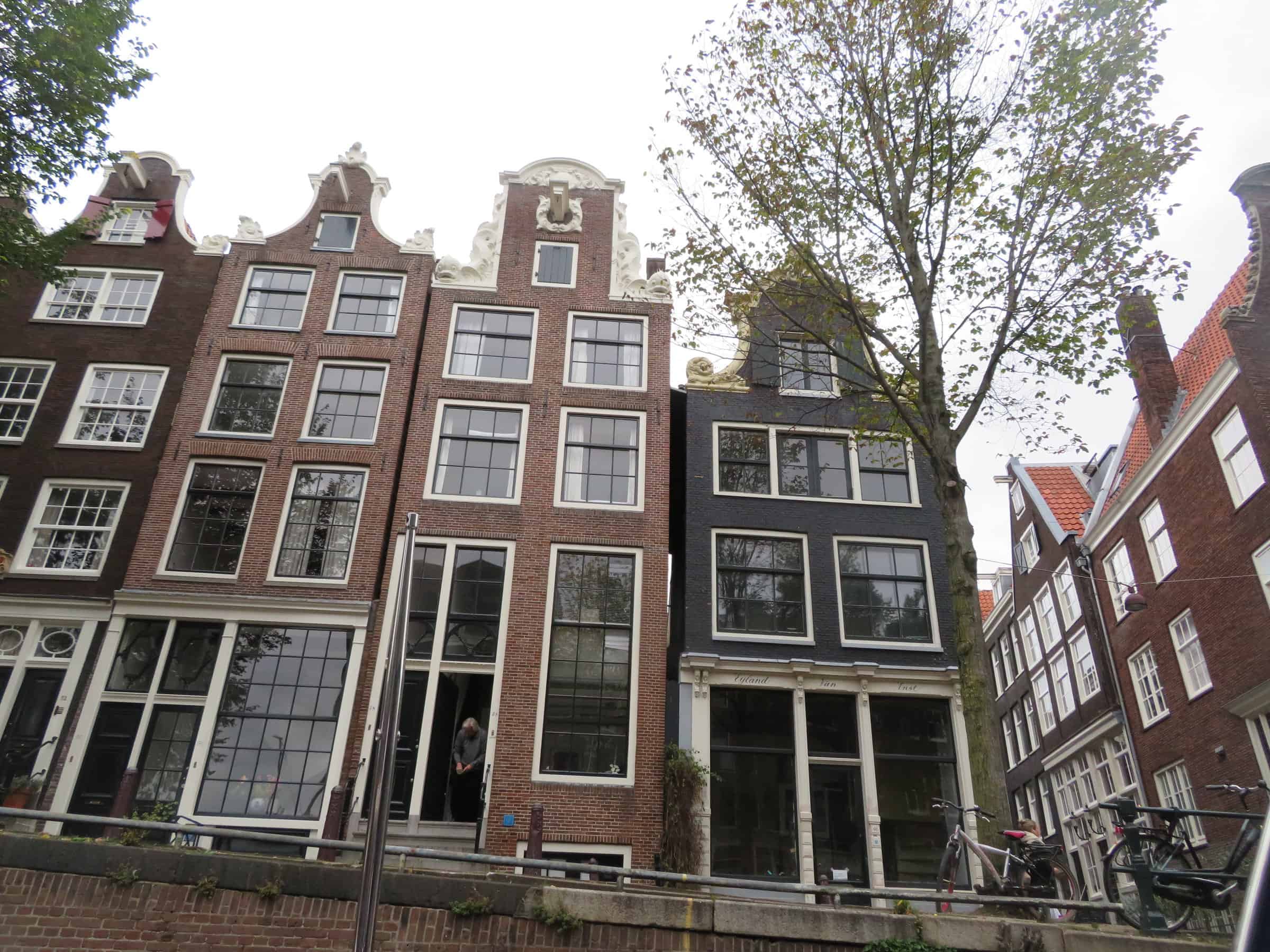 A row of traditional Dutch canal houses in Amsterdam, Netherlands, with their distinctive narrow facades and large windows. The buildings are adorned with ornate gables and sit alongside a canal, with bicycles parked nearby and a tree adding a touch of greenery to the urban scene. The overcast sky highlights the historic charm of this picturesque neighborhood.