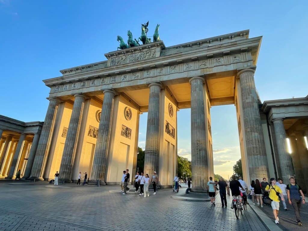 The iconic Brandenburg Gate in Berlin, Germany, stands tall under a clear evening sky. Sunlight bathes the neoclassical columns in a warm glow, while people stroll and cycle through the historic landmark. The gate, topped with a quadriga statue, serves as a symbol of peace and unity.