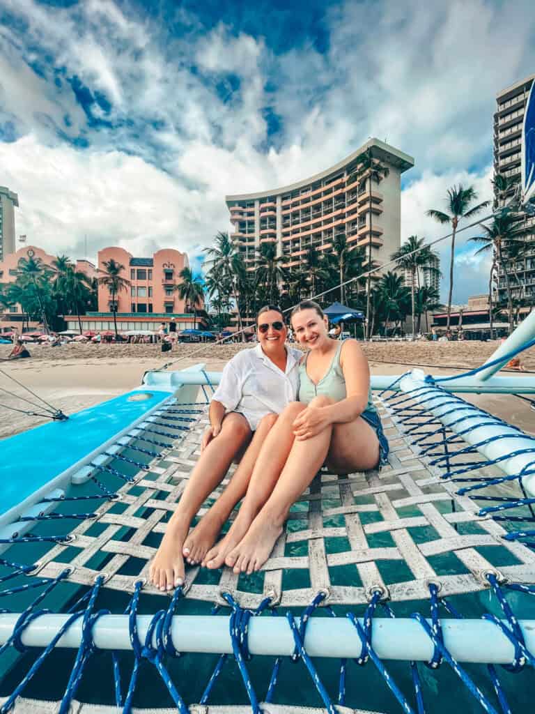 Two women smile while sitting on the net of a catamaran, with tall buildings and palm trees in the background. The sky is partly cloudy, and they appear to be enjoying a sunny day near the shore.