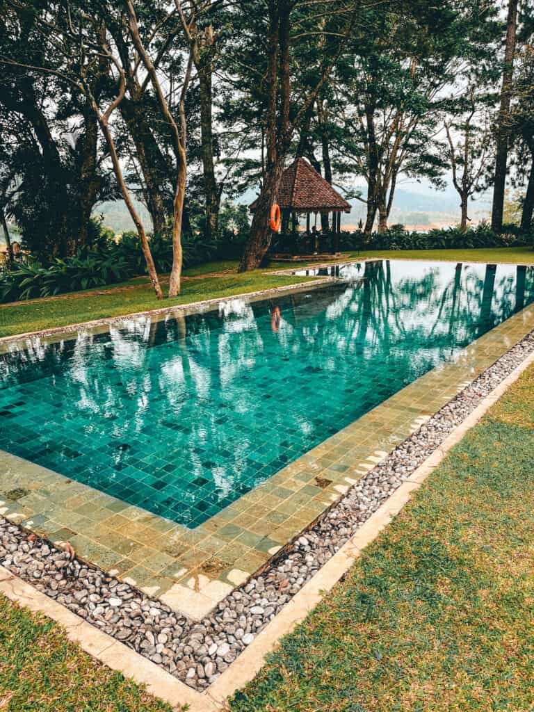 A tranquil outdoor swimming pool surrounded by green grass and trees, with a small pavilion in the background