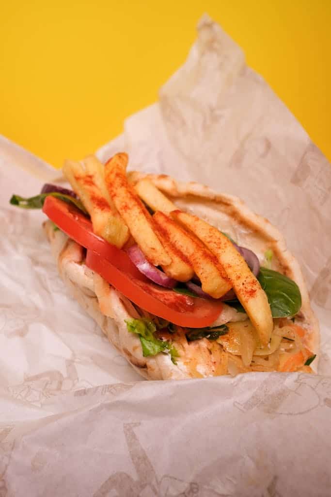 Delicious gyros with fried potatoes and sliced tomatoes with red onion and green leaves with cabbage leaves in pita on paper on yellow background