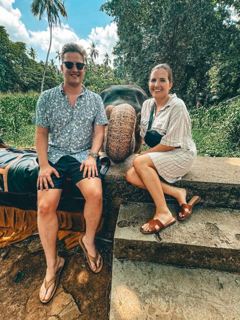 Two people sitting on a stone ledge next to an elephant's head, smiling at the camera. The man is wearing a blue patterned shirt, dark shorts, sunglasses, and flip-flops. The woman is wearing a striped dress, brown sandals, and a crossbody bag. The background features lush greenery and palm trees under a partly cloudy sky.