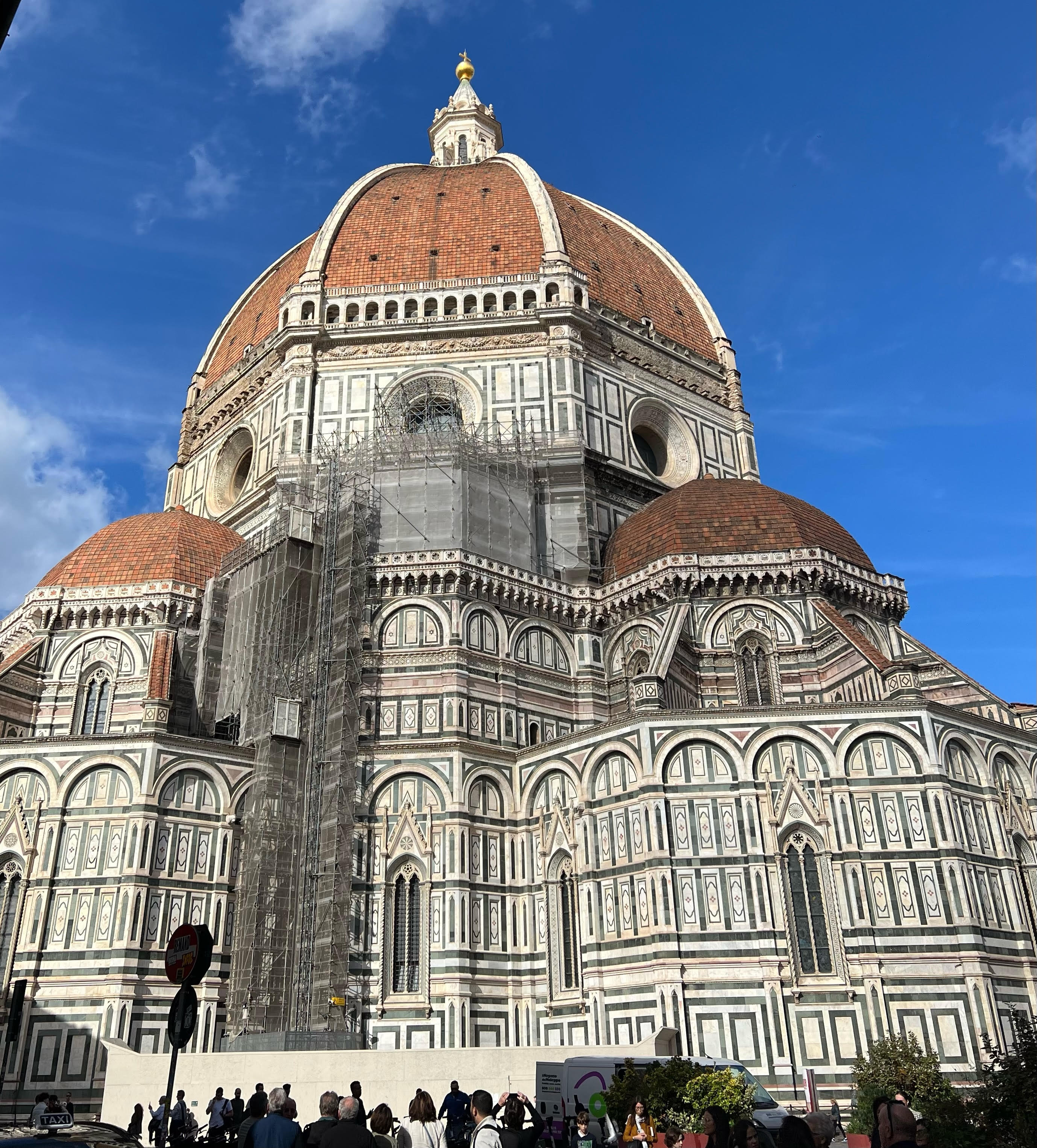 A detailed view of the Florence Cathedral, also known as the Duomo, in Florence, Italy. The magnificent red-tiled dome and intricate marble facade, featuring green, white, and pink panels, stand out under a clear blue sky. Scaffolding is visible on part of the structure, indicating ongoing restoration work, while visitors gather around the base, admiring the historic architecture.