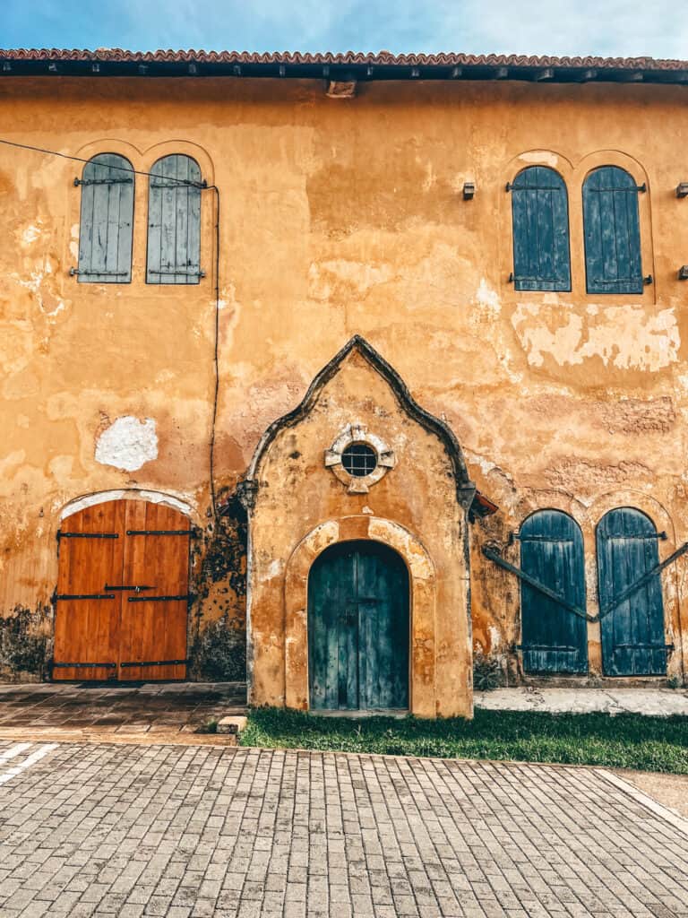 A historic building with weathered yellow walls, blue wooden shutters, and arched doorways, showing signs of age and history.