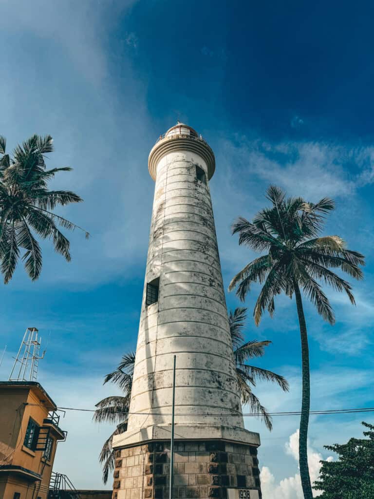 The Galle lighthouse standing tall with a white cylindrical structure, surrounded by palm trees and a bright blue sky