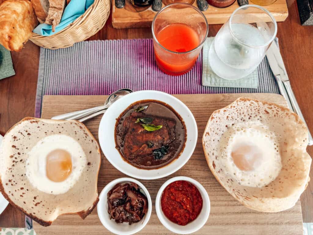 A traditional Sri Lankan breakfast featuring two egg hoppers, a bowl of curry, and side dishes including onion sambal and chili paste, served on a wooden board with drinks in the background.