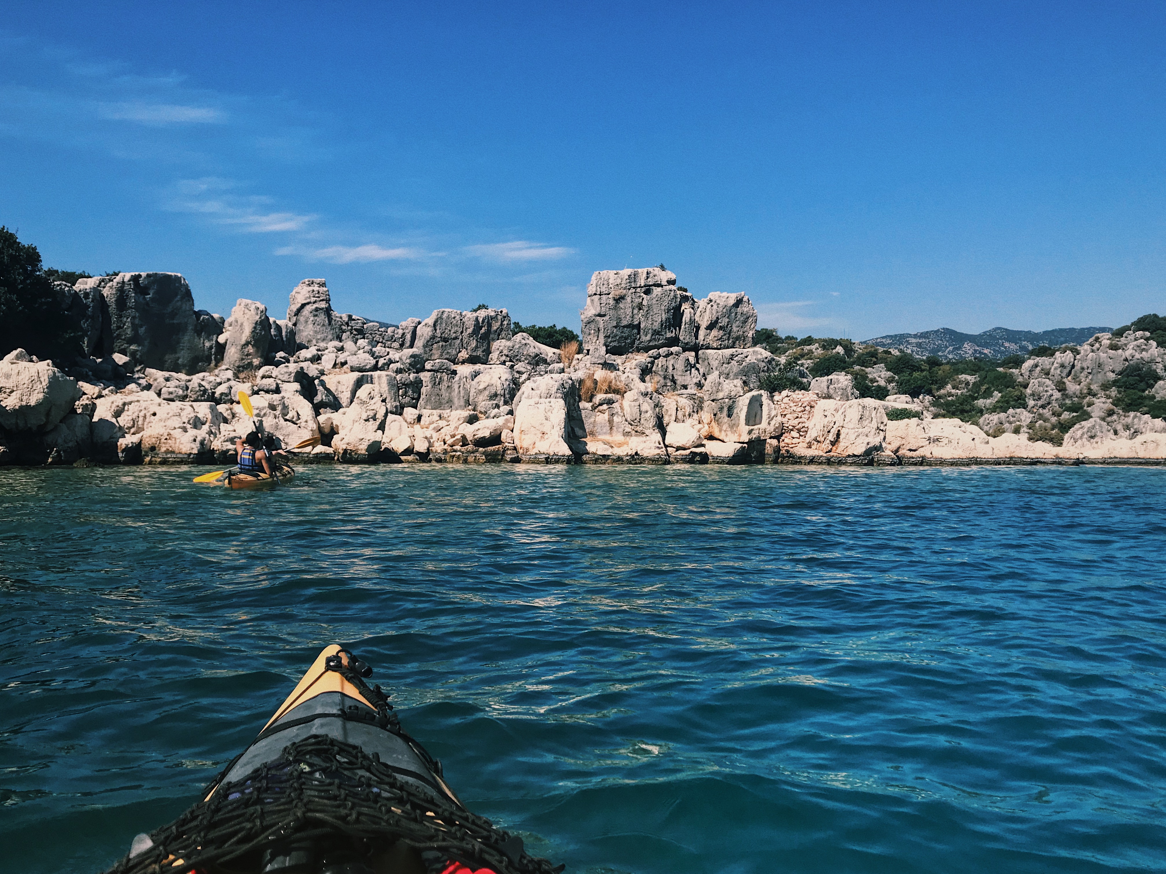 A kayaker paddles through the clear blue waters of Kaş, Turkey, approaching a rugged shoreline with rocky outcrops and sparse vegetation. The bright, sunny sky and distant mountains create a stunning backdrop, adding to the adventurous and scenic nature of this coastal destination. The perspective from another kayak emphasizes the immersive experience of exploring this beautiful landscape.
