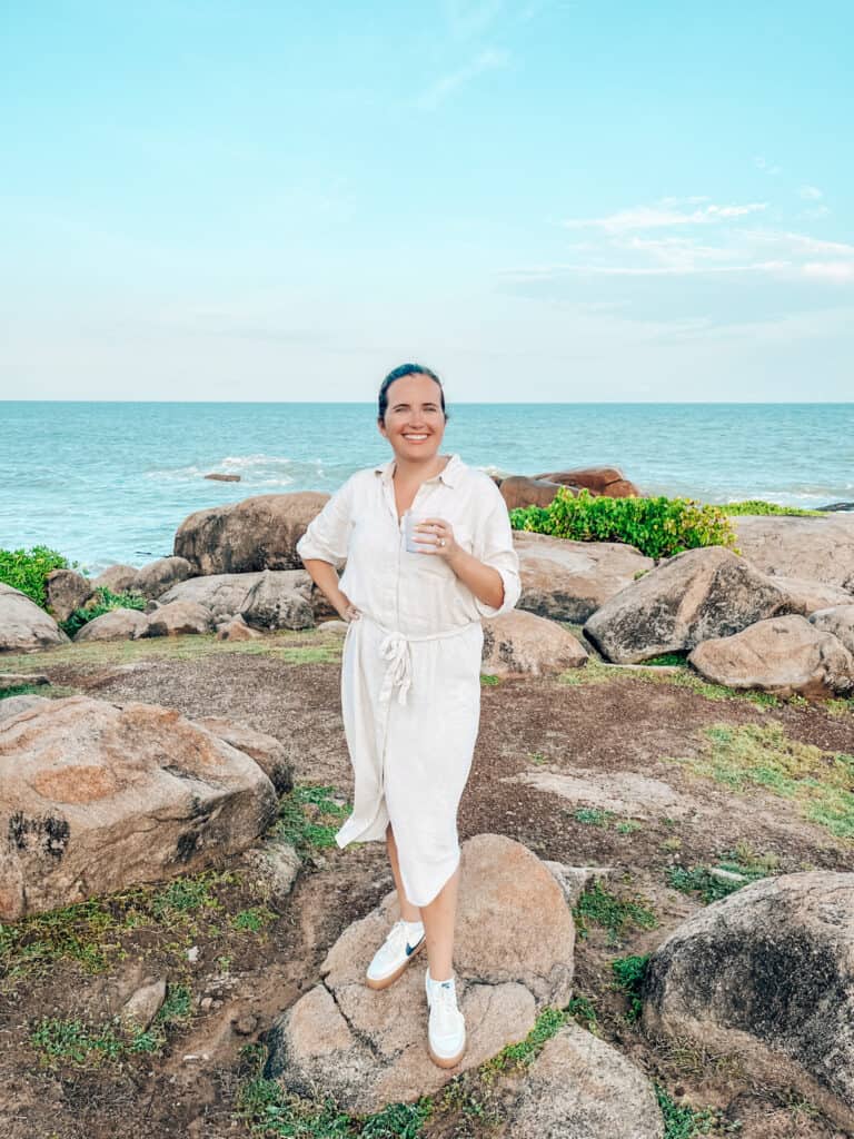 Person standing on rocky terrain by the ocean, wearing a white linen dress and white sneakers, holding a cup and smiling at the camera. The sky is clear with a few clouds, and the sea is calm in the background
