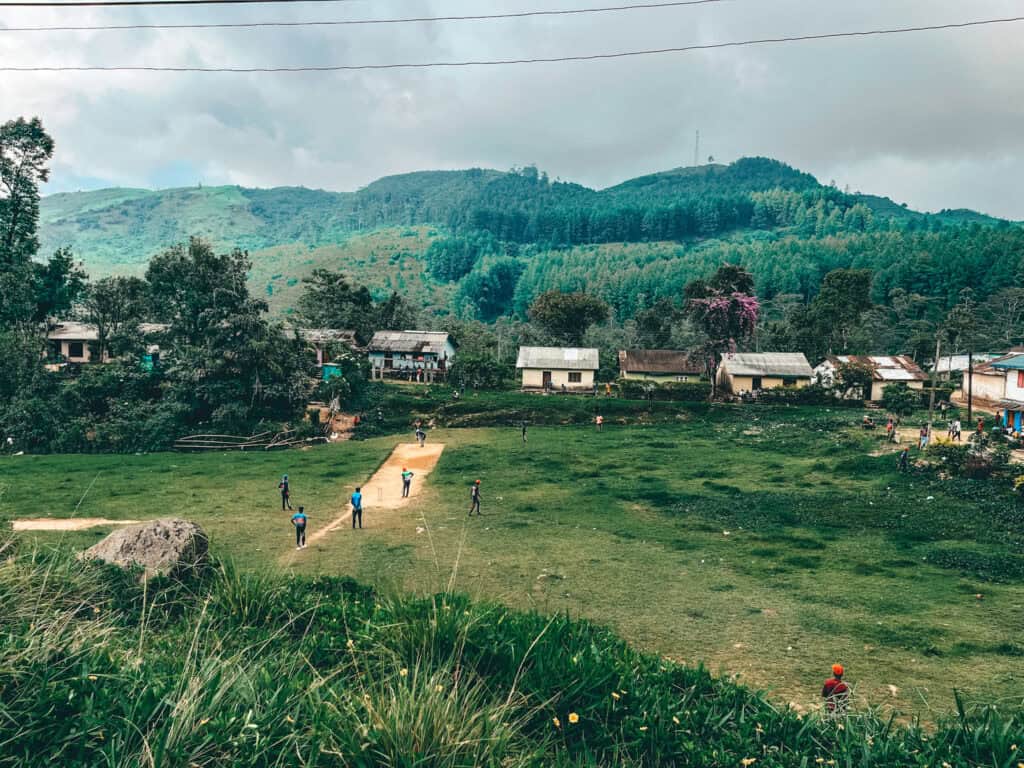 Children playing cricket on a grassy field in a local village in Sri Lanka, surrounded by small houses and lush green hills in the background
