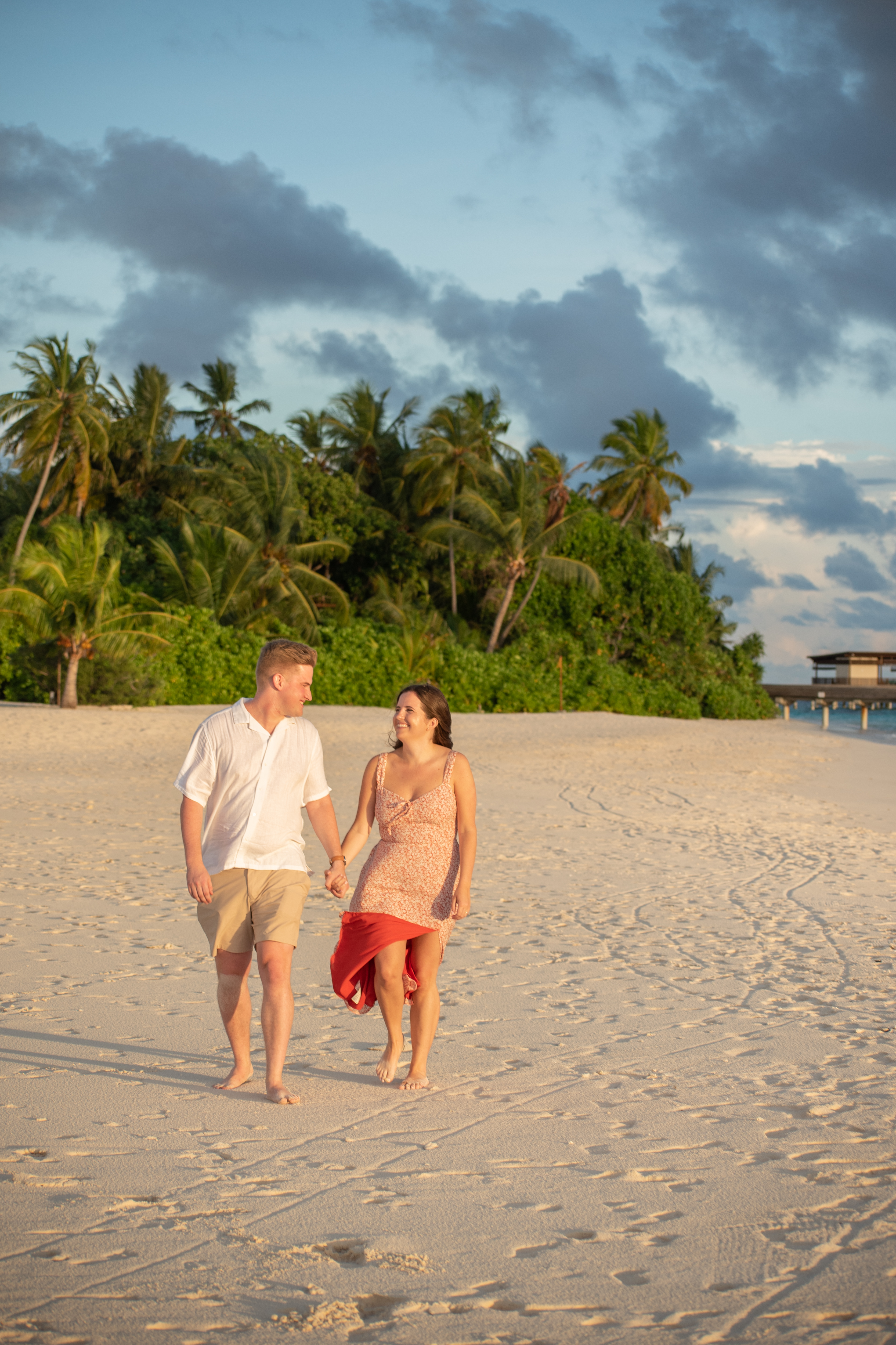 A couple walks hand-in-hand along a sandy beach at sunset, with lush palm trees and tropical foliage in the background. The woman wears a floral dress and the man a white shirt and beige shorts, both smiling as they enjoy their stroll.