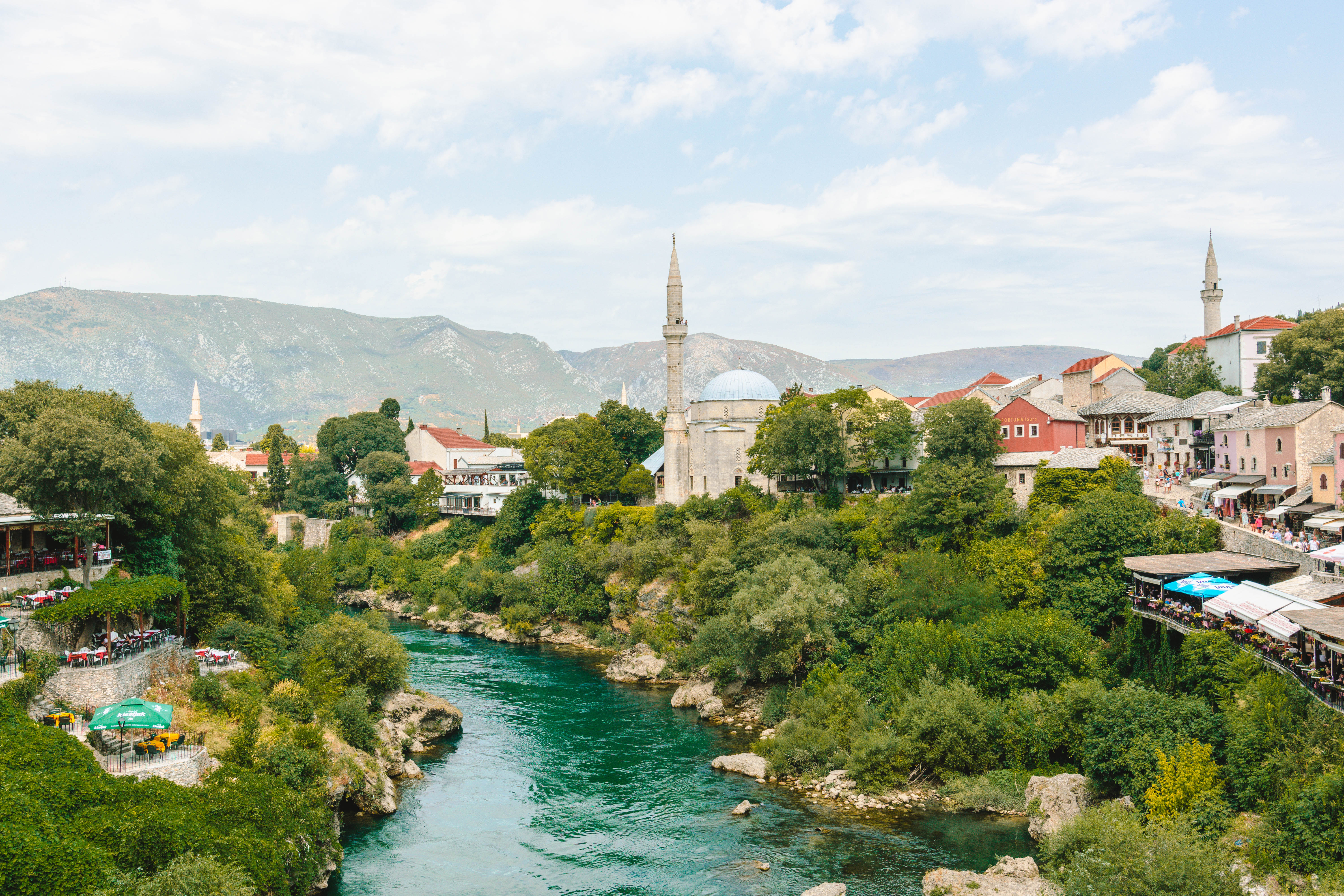 A scenic view of Mostar, Bosnia and Herzegovina, showcasing the Neretva River winding through the town. The landscape is dotted with historic buildings, including a prominent mosque with a tall minaret and a dome. The lush greenery along the riverbank and the surrounding hills create a picturesque setting under a partly cloudy sky.