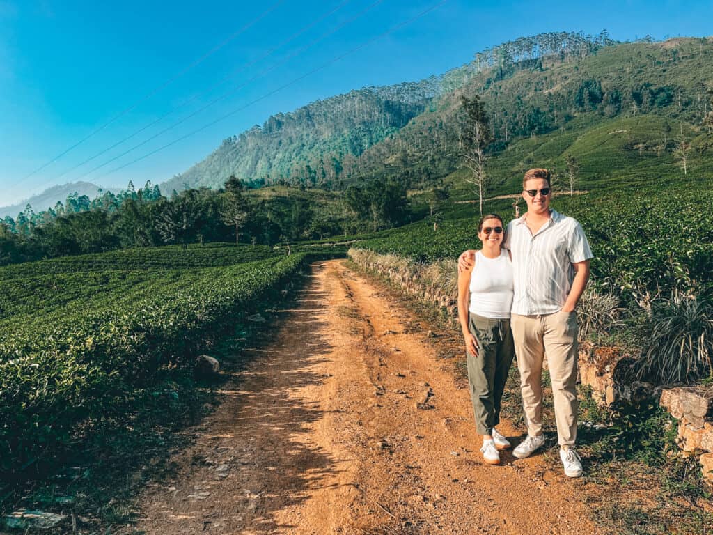 Two people standing on a dirt path in a lush green tea plantation with mountains in the background. The woman is wearing a white tank top, olive green pants, and white sneakers, while the man is wearing a striped short-sleeve shirt, beige pants, and white sneakers. Both are smiling and wearing sunglasses