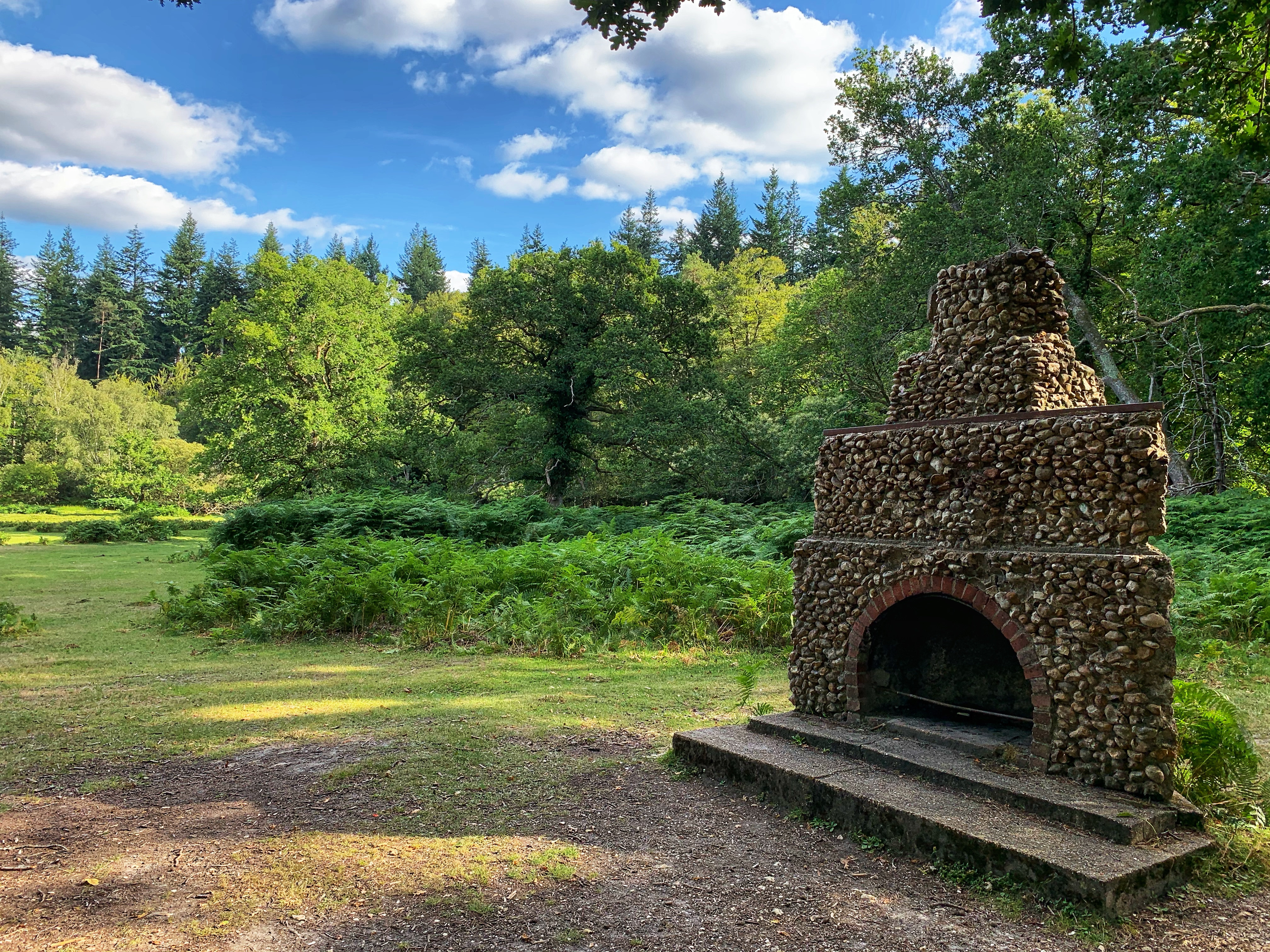 A serene scene in the New Forest, UK, featuring a rustic stone fireplace surrounded by lush greenery and dense woodland. The vibrant foliage under a bright blue sky with scattered clouds creates a peaceful and inviting atmosphere. The open grassy area and tall trees add to the natural beauty of this tranquil setting.