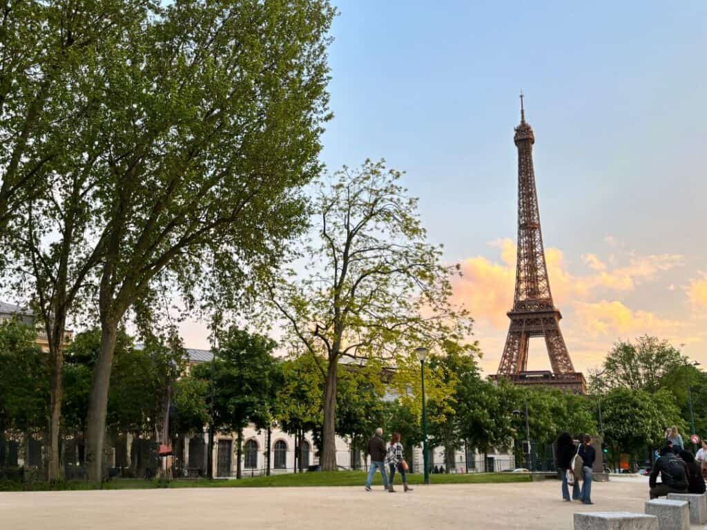 A view of the Eiffel Tower in Paris, France, at sunset, framed by lush green trees in a nearby park. People are gathered, enjoying the scenic spot and taking photos, while the sky glows with warm hues of pink and orange. The iconic landmark stands tall, adding a touch of elegance to the serene evening atmosphere.