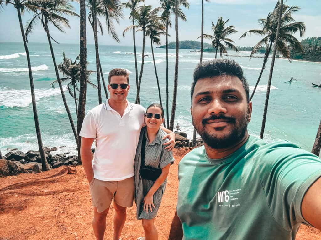 A group of three people, including a private driver, smiling and taking a selfie with tall palm trees and a turquoise ocean in the background.