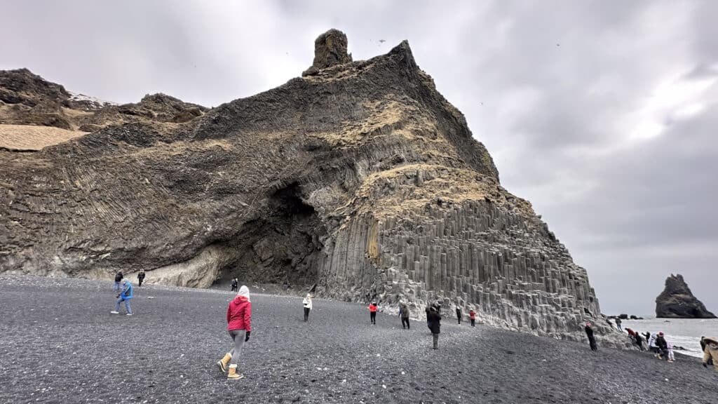 A dramatic landscape at Reynisfjara black sand beach in Vík, Iceland, featuring towering basalt columns and a rugged cliff under a cloudy sky. Visitors walk along the dark volcanic sand, exploring the unique geological formations and the cave nestled in the cliffside. The scene captures the stark, natural beauty of this iconic Icelandic location.