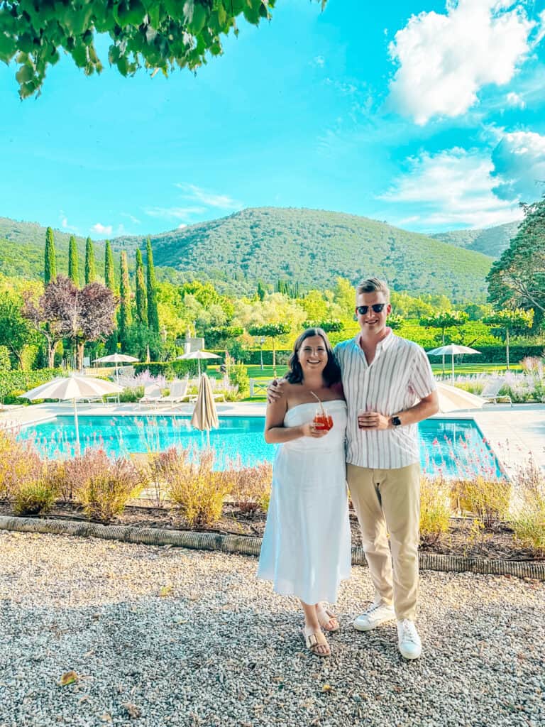 Couple wearing white standing in front of a bright blue pool, green trees, and green hills in tuscany italy
