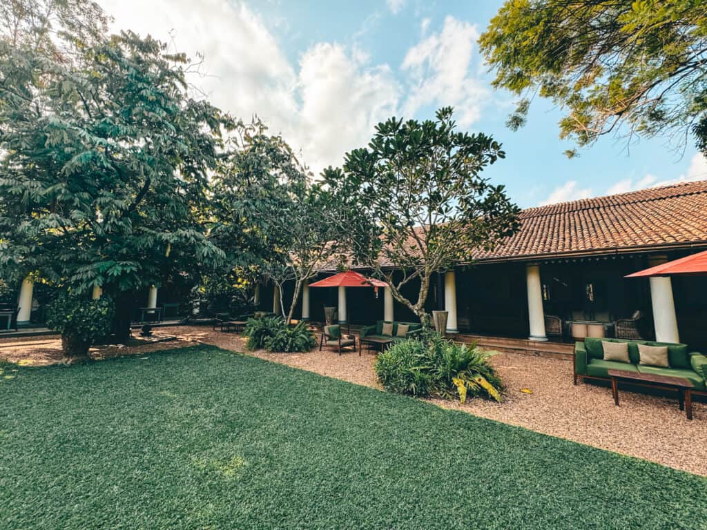 A serene courtyard featuring lush green trees and plants, outdoor seating with green cushions, and a building with a terracotta roof in the background