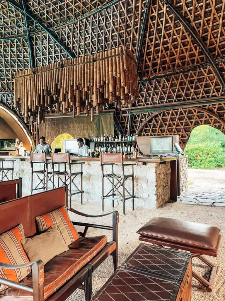 A cozy bar area with a bamboo chandelier hanging from the ceiling, high stools at a stone bar, and leather seating, all under a high, lattice-patterned roof.