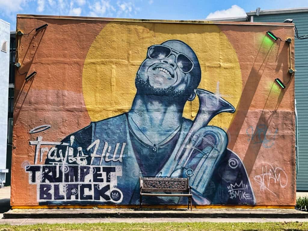 A vibrant mural in New Orleans, Louisiana, depicting musician Travis Hill, also known as Trumpet Black, joyfully playing his trumpet. The artwork, set against a colorful backdrop with a bright yellow circle, includes the artist's name and stylized graffiti elements, capturing the lively spirit of the city's music culture.