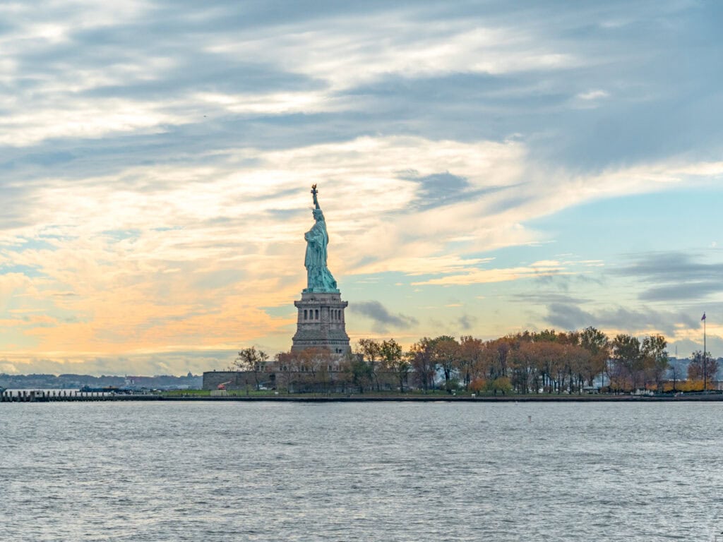 The Statue of Liberty standing proudly on Liberty Island in New York Harbor, framed by a dramatic sky with soft pastel clouds during sunset. The calm waters of the harbor lead to the iconic monument, surrounded by autumnal trees, symbolizing freedom and welcome.